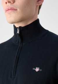 Navy blue zip-up sweater in a knit fabric with a high collar and embroidered logo on the left chest, featuring a silver zipper.