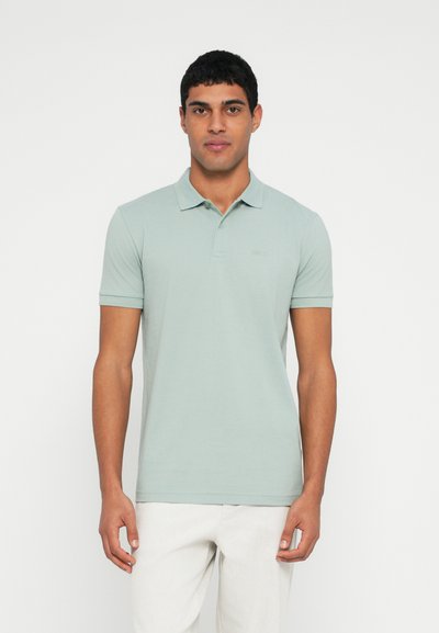 Polo vert clair avec une texture douce, manches courtes et col. Présente un logo discret sur la poitrine. Porté avec un pantalon de couleur claire.