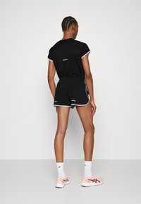 Schwarzes Sportoutfit mit kurzen Ärmeln und Shorts, mit weißen Akzenten. Modell trägt pinke Laufschuhe und weiße Knöchelsocken.