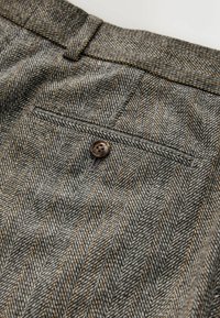 Gray herringbone-patterned fabric features a single back pocket with a brown button closure. The texture is woven and smooth.