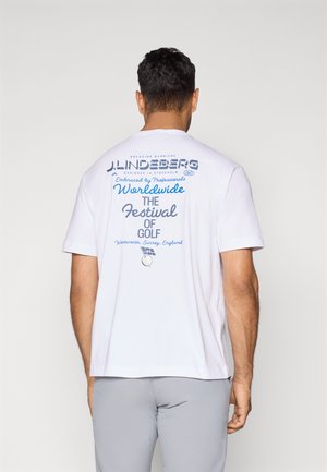 Férfi fehér pólót visel, melynek hátulján kék szöveg olvasható: "J. Lindeberg Worldwide The Festival of Golf Wentworth, Surrey, England".