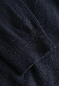 Tengerészkék kötött pulóver mandzsettája sima felületű, varrott logó részlettel. A csuklónál bordázott vége van, ami feszes illeszkedést biztosít.