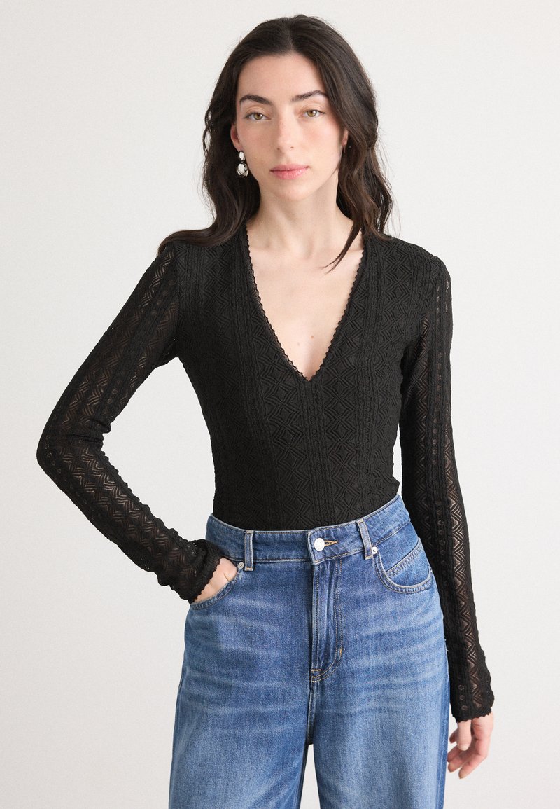 Musta pitsinen bodysuit, jossa on syvä V-pääntie ja pitkät hihat, ja jossa on geometrinen kuvio. Yhdistetty korkeavyötäröisiin sinisiin farkkuihin.