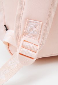 Batoh s růžovo-oranžovým popruhem vyrobeným z látky, s bílým logem, plastovou přezkou a tkanou texturou. Hladký, polstrovaný materiál.