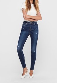 Jeans ajustados azul oscuro de material elástico, con detalles sutiles de desgaste y cintura alta, combinados con una blusa blanca y tacones blancos.