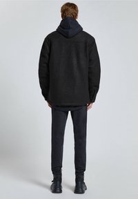 Giacca in pile nera con cappuccio, abbinata a pantaloni slim-fit scuri e sneakers nere, vista da dietro, caratterizzata da un design semplice.