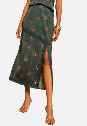 Femme portant une jupe midi verte fleurie avec une fente latérale, des bracelets dorés et des sandales à talons ouvertes dorées, debout devant un fond uni.