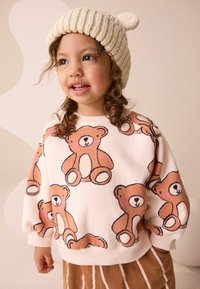 Vaaleanpunainen collegepaita, jossa on karhukuosi, pitkät hihat ja ribbireuna kauluksessa. Alla ruskeat raidalliset housut.