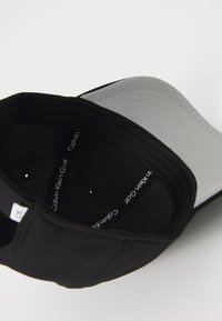 Μαύρο καπέλο με γκρι, υφασμάτινη βοήθεια. Ο εσωτερικός χώρος διαθέτει μαύρο ύφασμα με εκτυπωμένα λογότυπα "Calvin Klein Golf". Ορατές έξι τρύπες αερισμού.