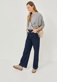 Denim wijd-uitlopende broek in donkerblauw, gecombineerd met een grijze gebreide trui en beige sneakers. Heeft een ontspannen silhouet en zijzakken.