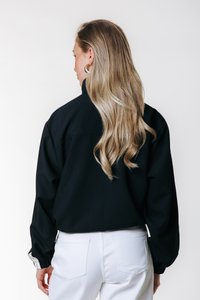 Chaqueta negra de gran tamaño con cuello alto y mangas largas abullonadas. Combinada con jeans blancos, que presentan un corte relajado con detalles de costura visibles.