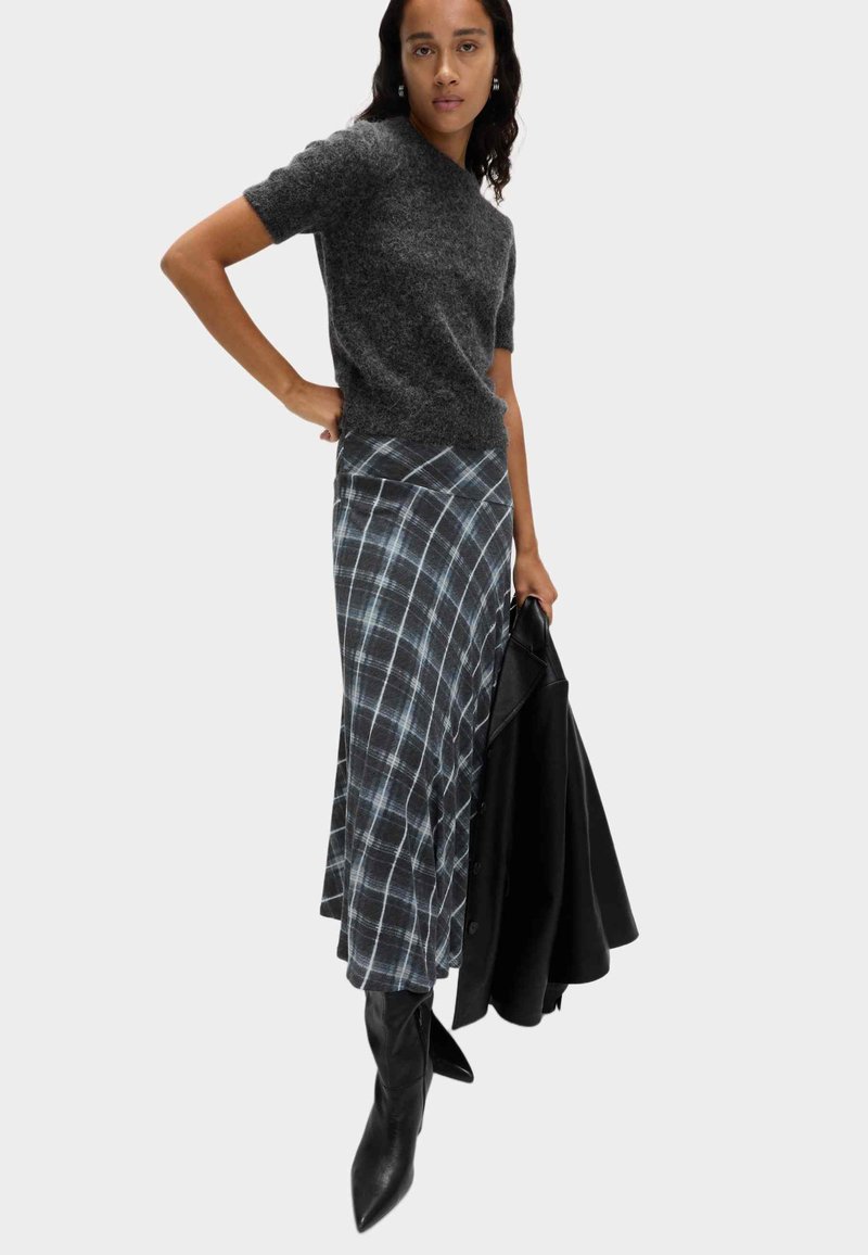 Grauer kurzärmliger Pullover kombiniert mit einem langen karierten Rock in Blau und Weiß, ergänzt durch schwarze kniehohe Stiefel und eine schwarze Jacke.
