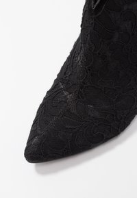 Escarpin noir en dentelle avec un design à bout pointu, arborant des motifs floraux et une surface texturée pour les détails.