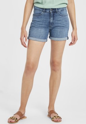 Person iført opkrullede blå cowboyshorts, lysegrøn top og brune flade sandaler med leopardprint, stående med afslappede arme.