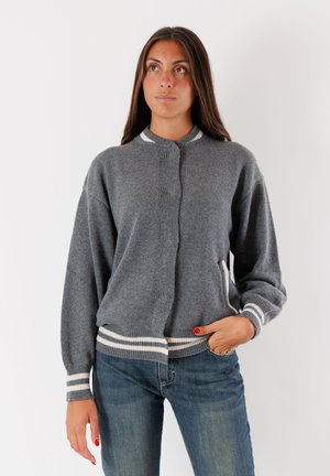 Donna che indossa un cardigan grigio a maglia con strisce bianche sul colletto, sui polsini e sull'orlo, abbinato a jeans blu, in piedi con una mano in tasca.