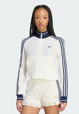 Mujer joven con trenzas largas que lleva una chaqueta de punto blanca con cremallera de Adidas y pantalones cortos a juego con rayas y logotipos azul marino.
