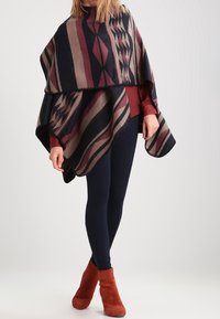 Poncho i ullblandat tyg med geometriska mönster i marinblått, vinrött och beige. Kanterna är mjukt fransade. Bärs över en vinröd topp.