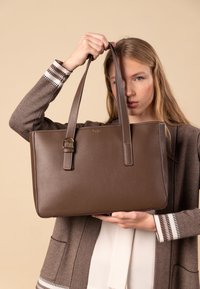 Brown leather tote bag with dual handles and a buckle accent. Smooth texture and structured shape, featuring a logo on the front.