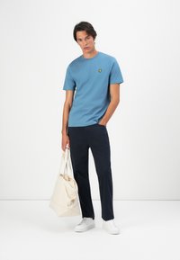 Tânăr îmbrăcat cu un tricou albastru cu logo, pantaloni negri, adidași albi, ținând o geantă mare bej, stând în fața unui fundal alb.
