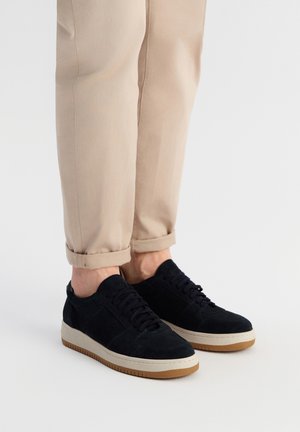 Black suede sneakers with laces and a beige rubber sole. Worn with rolled-up light beige trousers, showing the shoe's design and material.