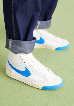 Białe sneakersy typu high-top z niebieskim znakiem i czubkiem, noszone z podwiniętymi ciemnymi jeansami na jasnizeleonej podłodze.