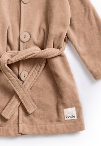 Beige bomullsrock med en texturerad tyg, stora knappar, knytning i midjan och ett märkesetikett med "Elodie" i svart.