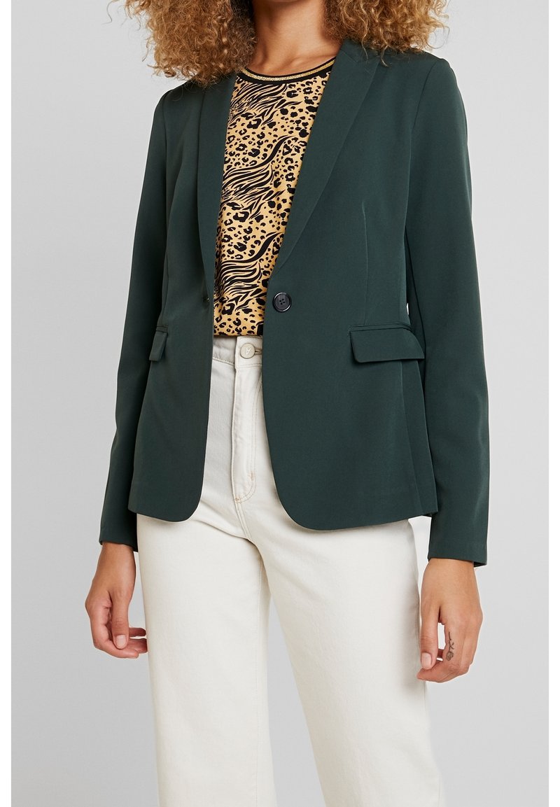 Femme portant un blazer vert foncé sur un haut imprimé léopard, associé à un pantalon blanc taille haute, se tenant devant un fond uni.