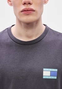 T-shirt cinzenta de algodão com decote redondo. Apresenta um pequeno patch com o logótipo "TOMMY JEANS" em azul e branco na área do peito esquerdo.