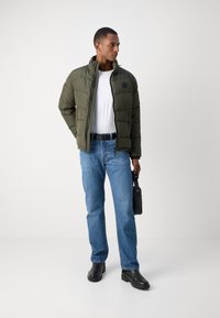 Olive green puffer jacket with a high collar, quilted texture, worn over a white t-shirt, paired with blue jeans and black boots.