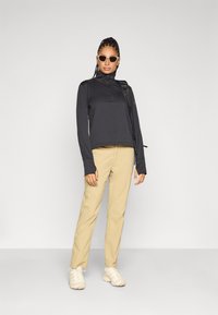 Μαύρο μακρυμάνικο τοπ με φερμουάρ στον γιακά, μπεζ παντελόνι και ανοιχτόχρωμα αθλητικά παπούτσια· ολοκληρωμένο με γυαλιά ηλίου και σακίδιο πλάτης.