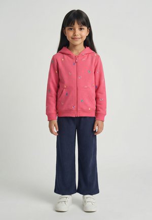 Jong meisje met lang donker haar dat een roze zip-up hoodie met kleurrijke patronen draagt, marineblauwe wijde broek en witte sneakers.