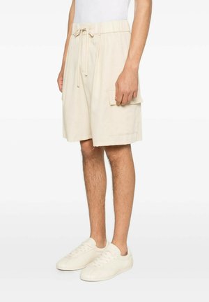 Uomo che indossa pantaloncini cargo beige al ginocchio con coulisse in vita e sneakers bianche, in piedi davanti a uno sfondo semplice.