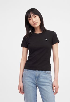 Giovane donna con capelli neri lisci che indossa una maglietta nera aderente a maniche corte e jeans azzurri, in piedi davanti a uno sfondo bianco.