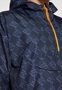Cortaviento azul marino con patrón de logotipos repetido, bolsillo frontal, capucha ajustable y detalles en cremallera de color naranja brillante. Ligero, con textura suave.