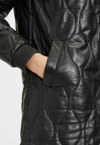 Cappotto in pelle nera con motivo trapuntato e polsini a coste. Dotato di una tasca laterale e di una texture liscia.