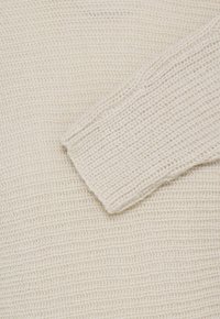 Gros plan sur un tissu tricoté couleur crème avec une manche visible, présentant un motif gaufré texturé.