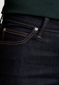 Mörkblå denimjeans med orange sömmar, som har en frampocket och metallhardware. Tyget har en mjuk textur med en subtil glans.