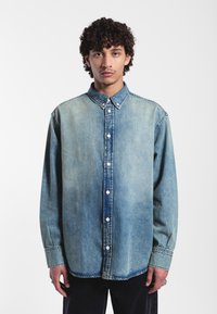 Camicia in denim azzurro chiaro con chiusura a bottoni, colletto a punta e maniche lunghe. Presenta una finitura sfumata e dettagli di cucitura visibili.