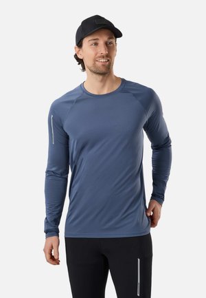 Mand iført en blå langærmet atletiktrøje og sort kasket, stående med hænderne nær hofterne, ser let mod højre og smiler.