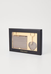 Tarjetero gris con ranuras para tarjetas y un llavero con un charm en forma de corazón, presentado en una caja de regalo negra con acentos dorados.