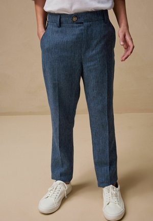 Person trägt blaue maßgeschneiderte Hose und weiße Sneaker, steht vor einem einfarbigen beigen Hintergrund mit entspannt an den Seiten hängenden Händen.