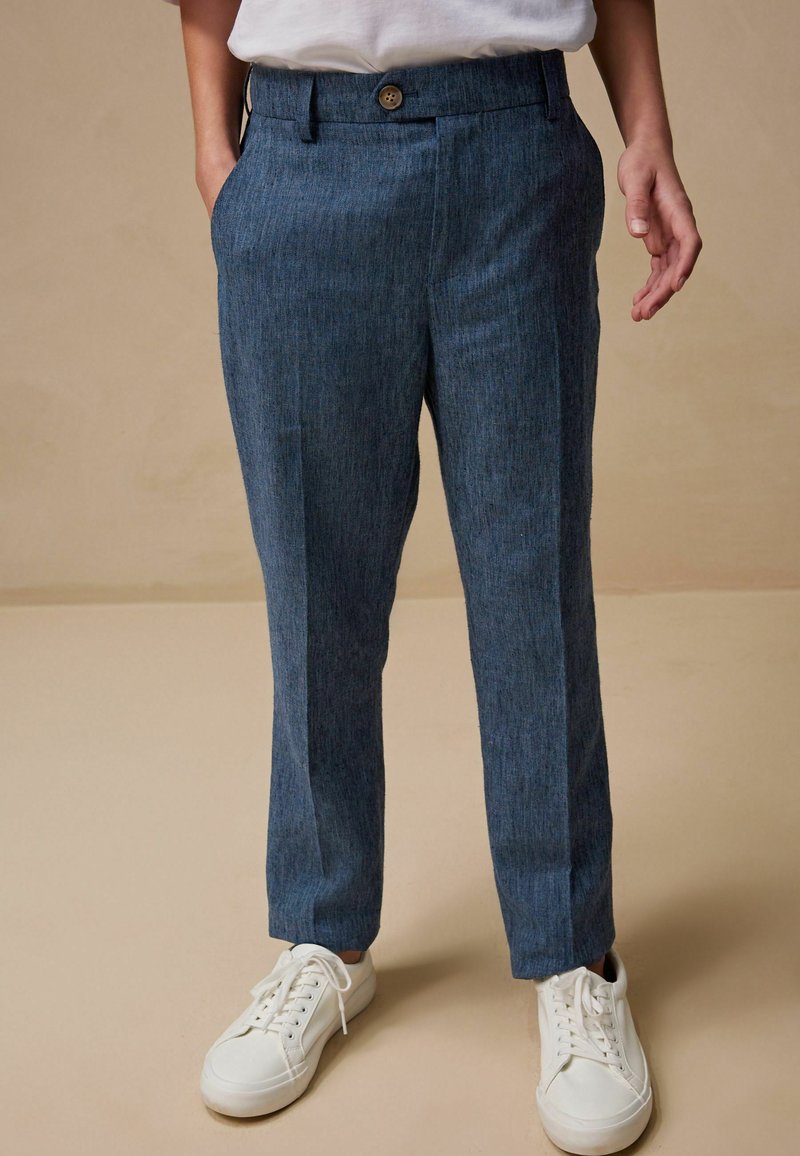 Persona che indossa pantaloni blu su misura e sneakers bianche, in piedi davanti a uno sfondo beige uniforme con le mani rilassate ai lati.