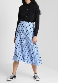 Chemise noire à manches longues, jupe plissée bleu clair avec des pois noirs, et baskets noires. La jupe possède une ceinture élastique pour le confort.