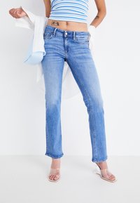 Jeans de denim azul claro de pierna recta con un ligero desgaste, diseño de cinco bolsillos y costuras en contraste. Combinados con un top corto a rayas y sandalias transparentes.