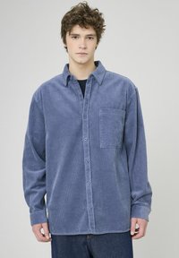 Chemise boutonnée en velours côtelé bleu avec une poche sur la poitrine, des manches longues et un col classique. La texture est côtelée avec un léger aspect brillant.