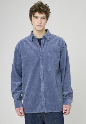 Chemise boutonnée en velours côtelé bleu avec une poche sur la poitrine, des manches longues et un col classique. La texture est côtelée avec un léger aspect brillant.