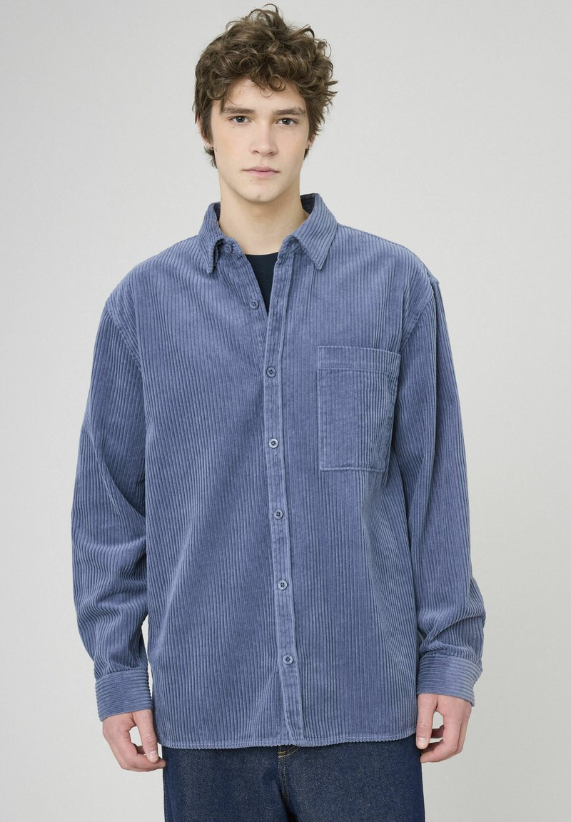 Chemise boutonnée en velours côtelé bleu avec une poche sur la poitrine, des manches longues et un col classique. La texture est côtelée avec un léger aspect brillant.