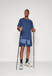 Tmavě modré tričko s jemným vzorem loga, doplněné modrými sportovními šortkami. Uživatel drží turistické hole, má na sobě světle šedé trailové boty a černé ponožky.