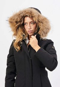 Donna che indossa un cappotto invernale nero con cappuccio foderato in pelliccia, che tiene il colletto con una mano, guardando leggermente di lato.