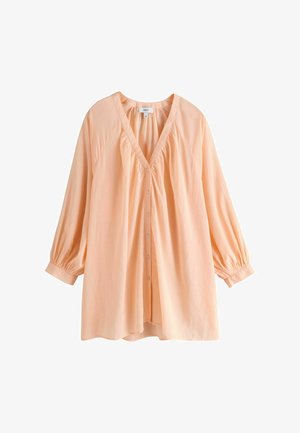 Femme debout en extérieur porte une robe ample couleur pêche claire avec de longues manches bouffantes et un collier blanc en perles grosses.